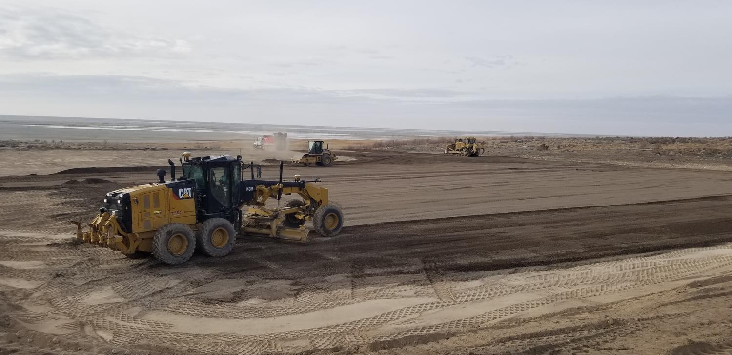 GPS-guided motor grader finish grading a large pad with a roller and dozer working behind